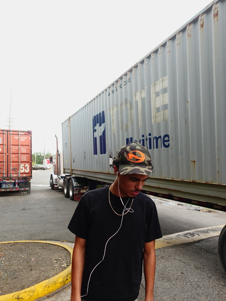 Model wearing the ALKOM distressed camo cap in an industrial loading yard.